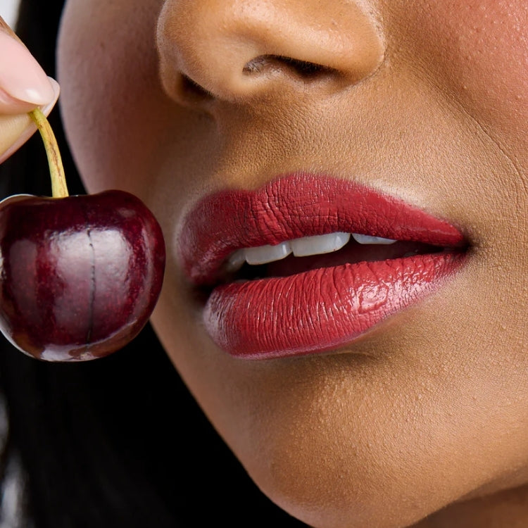Glaze High Colour Liquid Lipstick in een rijke bordeauxrode tint, aangebracht op volle lippen, met een kers als accessoire.